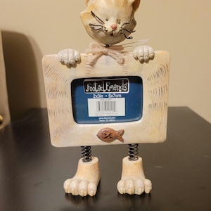 May include: A beige cat-shaped picture frame with a textured finish, featuring a head at the top, two paws holding the frame, and two feet attached by metal springs. The frame holds a label reading Footed Friends 2x3in 5x7cm with a fish icon.