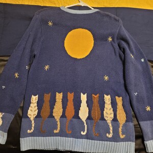 May include: A long-sleeved navy blue knit sweater featuring a large yellow moon and embroidered stars across the upper portion. The lower section displays a row of eight cats in various shades of brown, tan, and white, all facing away from the viewer.