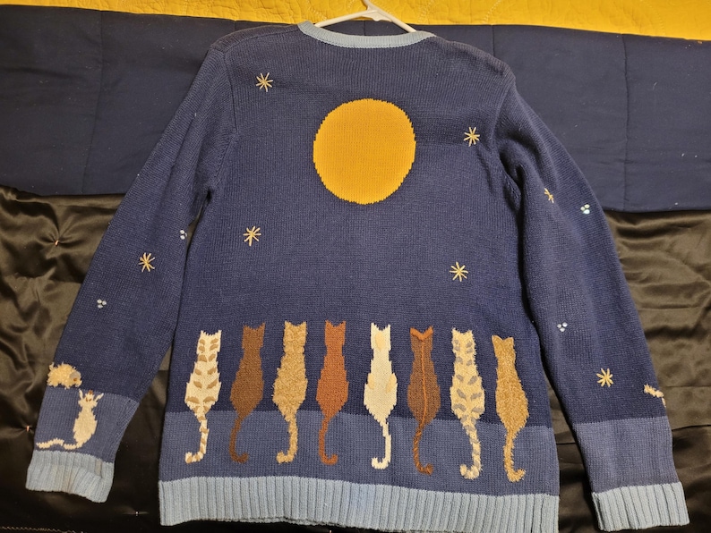 May include: A long-sleeved navy blue knit sweater featuring a large yellow moon and embroidered stars across the upper portion. The lower section displays a row of eight cats in various shades of brown, tan, and white, all facing away from the viewer.