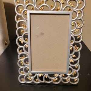 May include: Silver and gold decorative picture frame. The frame has a rectangular opening for a photo. The frame's border is silver with a repeating pattern of interlocking circles and gold leaf accents.