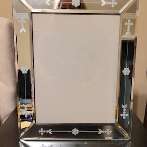 Puede incluir: Un marco de fotos rectangular espejado con dise&ntilde;os florales y geom&eacute;tricos grabados. El marco tiene un borde biselado y un centro de vidrio transparente para mostrar una fotograf&iacute;a o una obra de arte. El marco es plateado y negro.