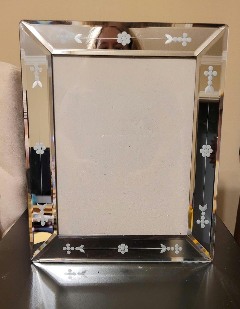 Puede incluir: Un marco de fotos rectangular espejado con dise&ntilde;os florales y geom&eacute;tricos grabados. El marco tiene un borde biselado y un centro de vidrio transparente para mostrar una fotograf&iacute;a o una obra de arte. El marco es plateado y negro.