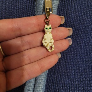 May include: A metal zipper pull featuring a small, cream-colored cat figurine with gold-toned stripes and bright green eyes. The cat charm is attached to a gold-colored zipper slider on a dark blue knitted garment with a light blue trim.