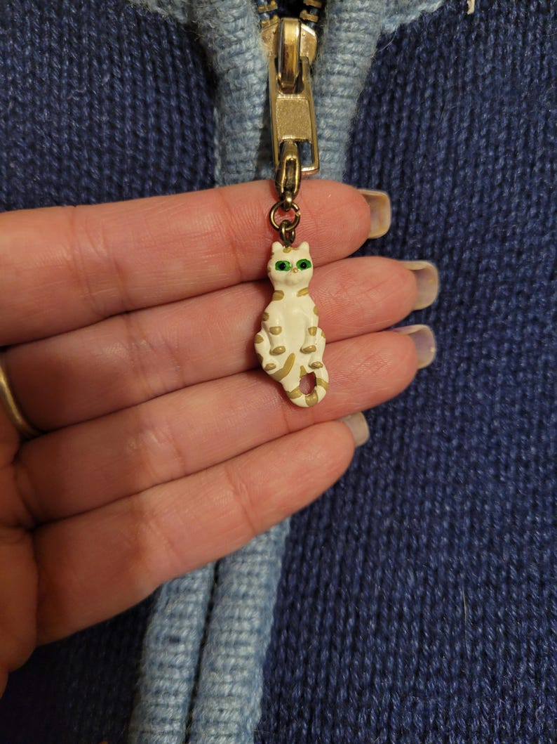 May include: A metal zipper pull featuring a small, cream-colored cat figurine with gold-toned stripes and bright green eyes. The cat charm is attached to a gold-colored zipper slider on a dark blue knitted garment with a light blue trim.