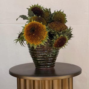 May include: A ceramic vase with a ribbed design holds a bouquet of sunflowers. The sunflowers have yellow and brown petals with green leaves. The vase is dark green and brown, and sits on a dark brown table.