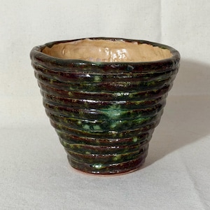 May include: A handmade ceramic pot with a textured, ridged exterior. The pot features a gradient of colors, including dark green, burgundy, and a light tan interior. The pot's shape is wider at the top and tapers towards the base.