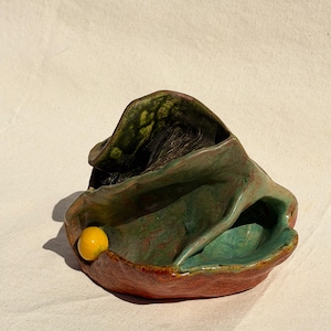 May include: A handcrafted ceramic bowl with a unique, organic shape. The bowl features a blend of colors, including green, red, and yellow. The interior has a dark, textured appearance, and a small yellow sphere is attached to the side.