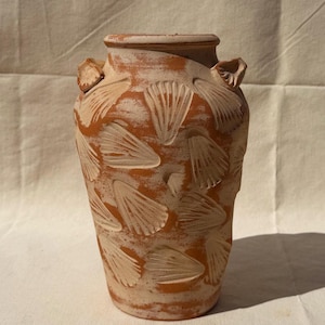 May include: A terracotta vase with a textured surface and a light brown color. The vase features a repeating seashell pattern carved into the surface. Two small handles are on the sides of the vase.