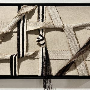 May include: Abstract textile art piece in a black frame. The artwork features woven ribbons in cream, black, and brown, creating a layered, textured design. A knotted detail and hanging fringe add visual interest to the neutral color palette.
