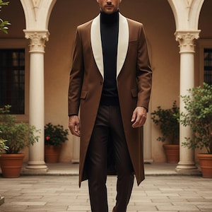 May include: A brown overcoat with a white lapel, paired with brown trousers and shoes. The outfit includes a black turtleneck sweater. The coat is long and tailored, creating a sophisticated look. The setting is an outdoor courtyard with stone flooring.