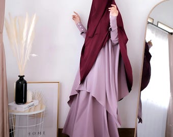 May include: A modest, layered dress in a soft lavender hue, paired with a deep burgundy hijab and niqab. The dress features long sleeves and a flowing skirt. The outfit is suitable for formal occasions.