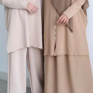 May include: Two individuals are shown wearing matching modest outfits. The outfits consist of long-sleeved tops with button closures and wide-leg pants or a skirt, all in neutral beige and tan tones. The outfits include head coverings.