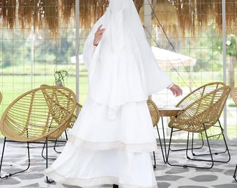 May include: A white maxi dress with a lace trim and a matching white niqab. The dress has a flowing skirt and a fitted bodice. The woman is wearing a gold crown.