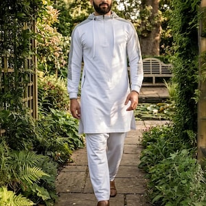 May include: A white hooded tunic and matching pants set with gray accents on the sleeves and hood. The tunic has a quarter-zip closure. The outfit is worn with brown shoes. The person is walking on a stone path in a garden.