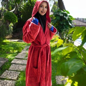 May include: A red hooded bathrobe with a matching belt and pockets. The robe is made of a soft, plush material. The person is wearing a black hijab and blue gloves. The background features a garden with green foliage and a stone pathway.