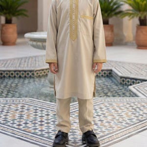 May include: A beige two-piece outfit for children. The long-sleeved tunic features gold embroidery around the collar, down the front, and on the cuffs. Matching pants and black shoes complete the ensemble. The outfit is suitable for special occasions.