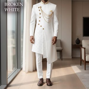 Puede incluir: Un traje blanco roto con detalles dorados. La chaqueta presenta botones dorados, una cadena decorativa y un diseño asimétrico. Los pantalones son rectos y el conjunto se completa con zapatos marrones. El texto "BROKEN WHITE" es visible.