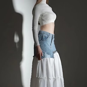 May include: A white crop top with long sleeves and a denim and white maxi skirt. The skirt has a denim panel at the waist and a tiered, flowing white fabric. The model is wearing clear high heels.