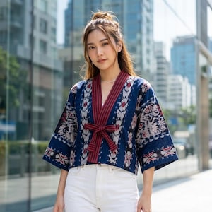 May include: A blue and white patterned kimono-style jacket with a burgundy trim and bow. The jacket has short sleeves and is paired with white pants. The background features a modern cityscape with glass buildings.