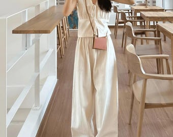 May include: A sleeveless cream-colored outfit consisting of a top and wide-leg pants. The top has a V-neck and tie detail, paired with a small pink crossbody bag and silver sandals. The outfit is simple and elegant, suitable for casual wear.