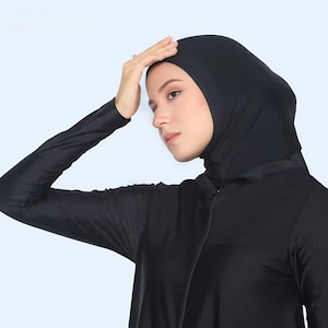 Puede incluir: Un traje de baño negro de manga larga con capucha. El traje de baño tiene una cremallera completa y una textura acanalada en el torso y las mangas. La modelo lleva el traje de baño y tiene la mano en la frente.