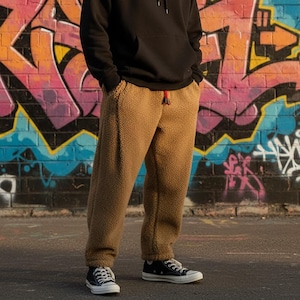 May include: A person wearing a black hoodie and tan fleece sweatpants with a red drawstring. The person is wearing black and white sneakers. The background is a colorful graffiti wall.