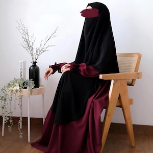 May include: A black and burgundy abaya, a traditional garment, is worn by a person seated in a wooden chair. The abaya features a black head covering and sleeves with burgundy accents. The lower portion of the garment is burgundy.