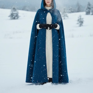May include: A hooded, long blue cape with a button closure, worn over a white knit sweater. The cape is knee-length and worn in a snowy outdoor setting. Black gloves are visible.