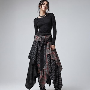 Puede incluir: Una blusa negra de manga larga combinada con una falda asimétrica de varias capas que mezcla tela negra con patrones de cuadros grises y rojos. La falda incluye un detalle de lazo en la cintura con paneles de tela colgantes y un dobladillo irregular.