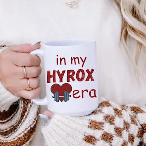 May include: White ceramic mug with the text "in my HYROX era" in red and black. The mug features a heart and dumbbell graphic. The mug is held by a person wearing a cream-colored sweater with brown and white checkered sleeves.