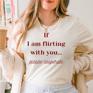 Może przedstawiać: Kremowa koszulka z napisem "If I am flirting with you... please cooperate" w kolorze bordowym. Osoba ma na sobie szare spodnie dresowe i kremowy kardigan. Napój kawowy trzymany jest w lewej ręce.