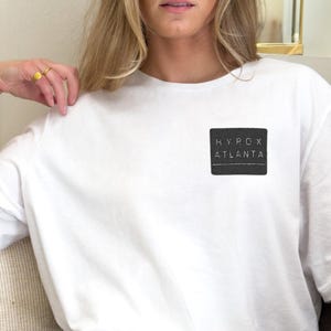 May include: White t-shirt with a black square patch that reads "HYPOX ATLANTA". The shirt is paired with brown trousers. The person is sitting on a sofa with cushions.