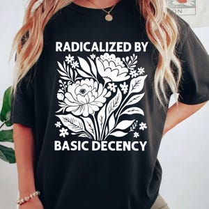 以下が含まれることがあります： 白い花柄と「RADICALIZED BY BASIC DECENCY」の文字が入った黒いTシャツ。 グラフィックは様式化された花と葉が特徴です。 シャツは柔らかい素材で作られています。