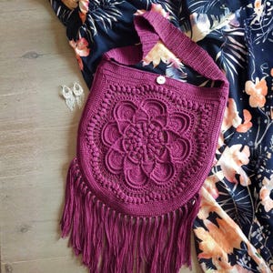May include: A deep magenta crochet shoulder bag featuring a central circular mandala-style pattern, a single wide strap, a small white button closure at the top, and long fringe detailing along the bottom edge of the bag.
