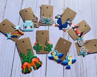 Op de afbeelding: Een verzameling van 10 paar pixel art oorbellen op bruin karton. De oorbellen hebben verschillende ontwerpen, waaronder een regenboog, een melkpak, een cactus, een vogel, een papegaaiduiker en een konijn.