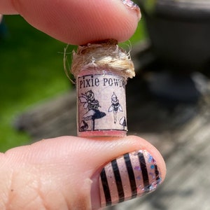 May include: A small glass bottle with a cork stopper and a label that reads "Pixie Powder". The label features a fairy with wings and a mushroom. The bottle is held in a hand with pink and black striped nail polish.