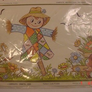 May include: A vintage craft pattern featuring a smiling scarecrow with a patchwork outfit, standing in a field with flowers and a fence. The pattern is titled "Carousel Crafts 1969" and has a pattern code of "P1000".