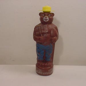 May include: A brown plastic figurine of a bear wearing a yellow hat and blue jeans. The bear is holding a shovel and a fire extinguisher. The figurine is a vintage Smokey Bear.