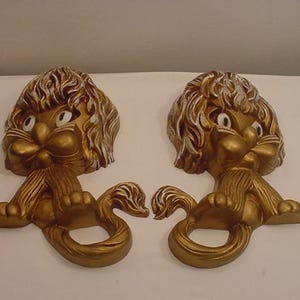 Puede incluir: Un par de esculturas de pared de león pintadas en oro con detalles en blanco. Los leones tienen ojos grandes y están sentados con sus colas enroscadas alrededor de sus patas.