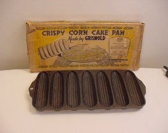 May include: Vintage Griswold cast iron corn cake pan in a box. The box is yellow with black text that reads "Crispy Corn Cake Pan Made by Griswold". The pan has six individual molds shaped like corn cobs.
