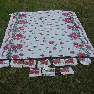 May include: A white tablecloth with a red and green poinsettia pattern. The tablecloth is folded and laid out on a green grassy surface. There are 14 matching napkins folded and laid out in a row below the tablecloth.