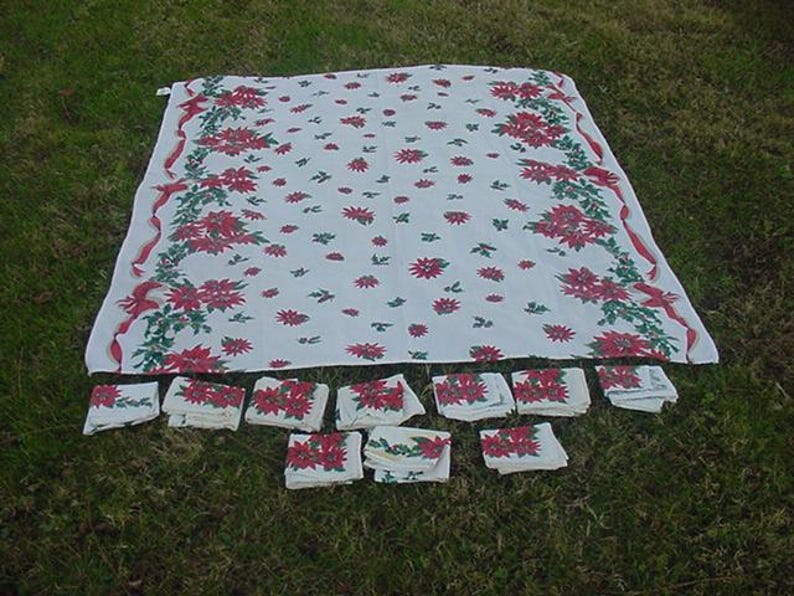 May include: A white tablecloth with a red and green poinsettia pattern. The tablecloth is folded and laid out on a green grassy surface. There are 14 matching napkins folded and laid out in a row below the tablecloth.