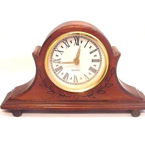 May include: A small, wooden quartz clock with a gold-colored face and Roman numerals. The clock has a curved top and a rectangular base with decorative leaf accents. The hands point to approximately 3:45.