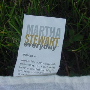 May include: A white fabric label with the text "MARTHA STEWART everyday" in gray and gold lettering. The label also includes the text "100% Cotton" and care instructions for machine washing and drying.