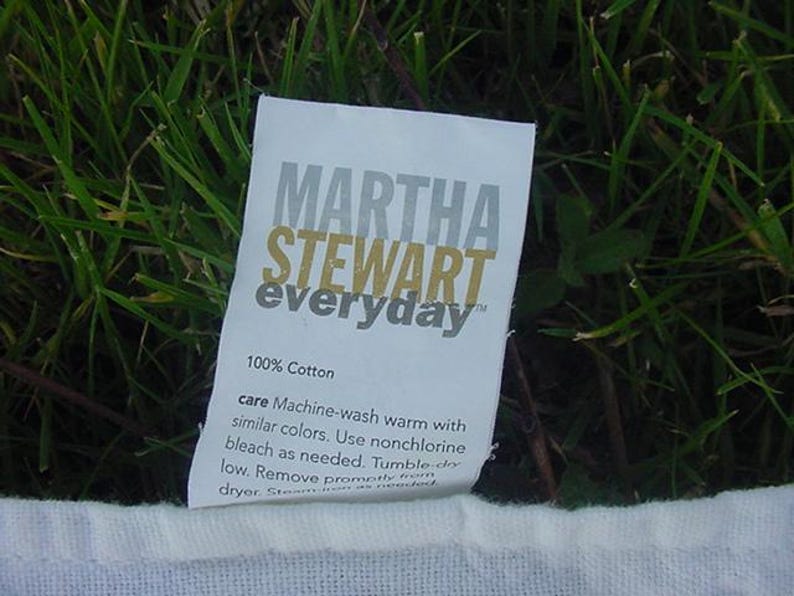 May include: A white fabric label with the text "MARTHA STEWART everyday" in gray and gold lettering. The label also includes the text "100% Cotton" and care instructions for machine washing and drying.