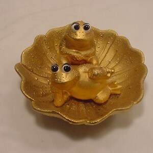 May include: Two gold frog figurines with black eyes sitting on a gold seashell-shaped dish.