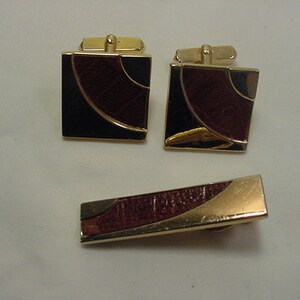 Może przedstawiać: Zestaw złotych spinek do mankietów i klipsa do krawata. Spinki do mankietów są kwadratowe z geometrycznym wzorem w kolorze czarnym i brązowym. Klips do krawata ma podobny wzór.