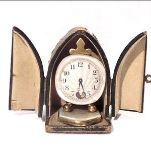 May include: A vintage travel clock with a white face and black Roman numerals. The clock is housed in a black leather case with a gold-colored clasp. The clock's face is approximately 2 inches in diameter.