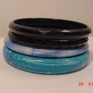 May include: A stack of four plastic bangles. The top bangle is black with a faceted design. The second bangle is dark blue with a smooth finish. The third bangle is light blue with a marbled pattern. The bottom bangle is turquoise with a marbled pattern.