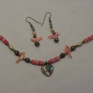 May include: A silver necklace with pink and white beads, a heart pendant, and two matching earrings with pink bird charms.